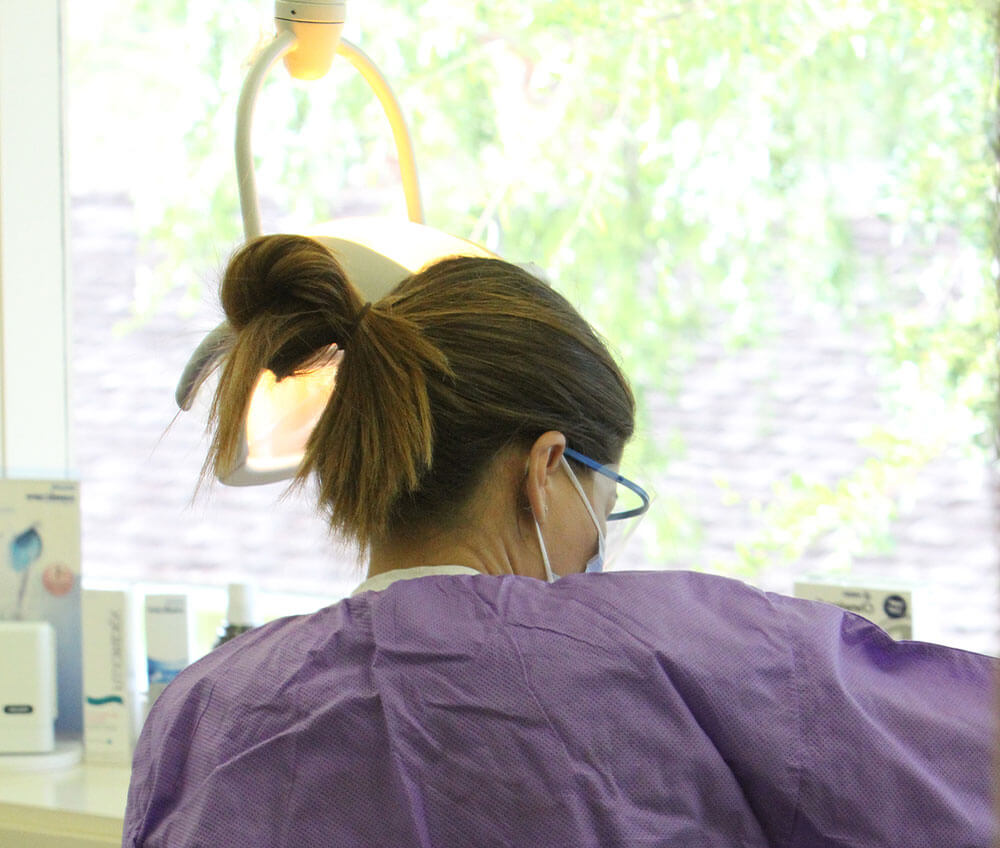 A dental assistant, in a purple gown, googles and face mask, with her back toward the camera