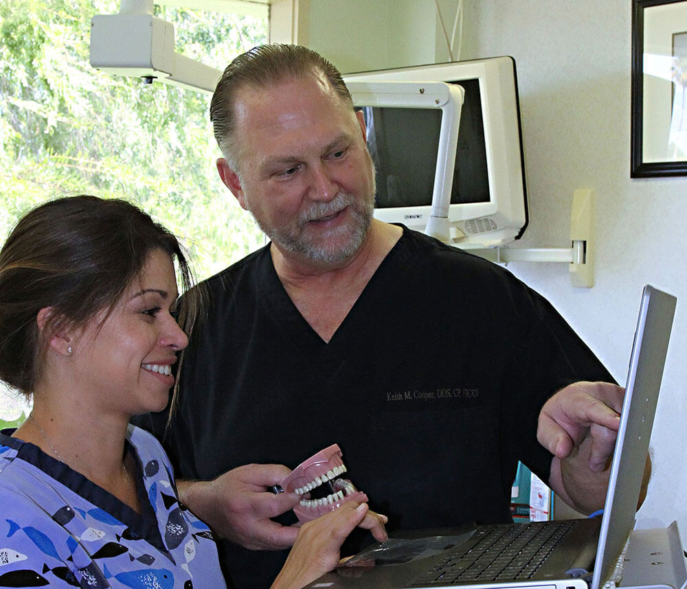 Dr. Cooper holds a model of teeth and points to the screen on a laptop, while a dental assistant observes