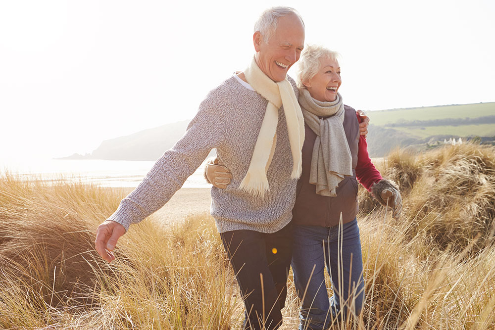 Older couple walking through tall grass with hills and a beach in the background